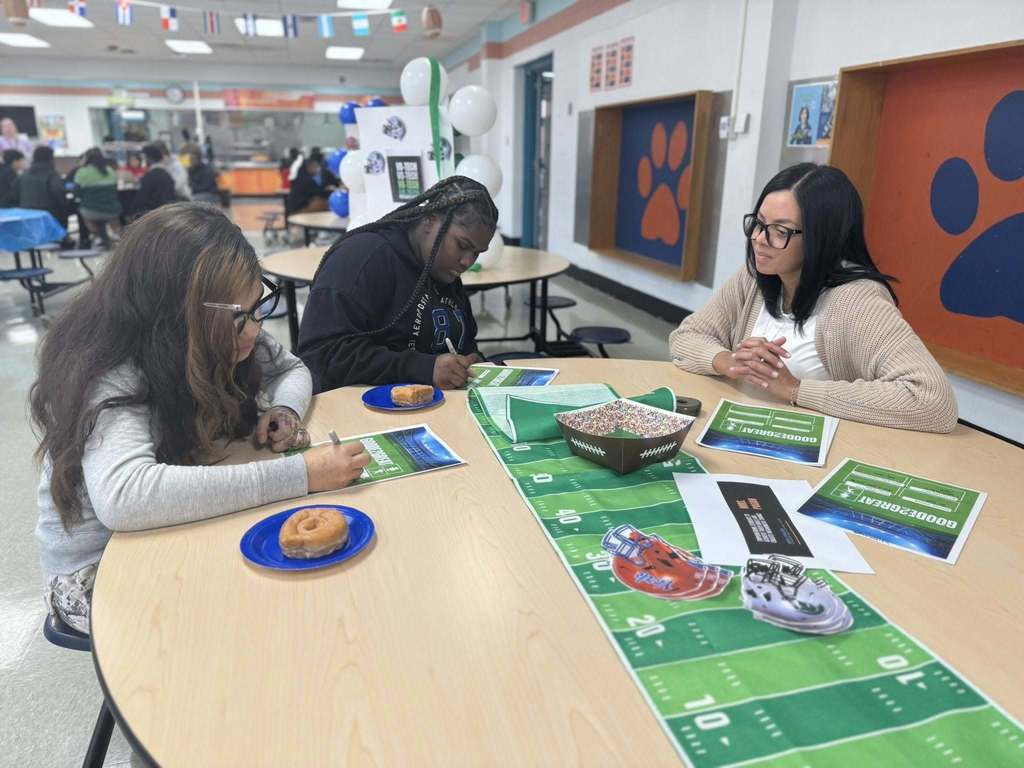 Two students and a district staff member are sitting at a table in a school cafeteria. Both of the students are writing on papers with a football field tablecloth and snacks nearby, while the district staff member is observing them. Other people can be seen in the background. 