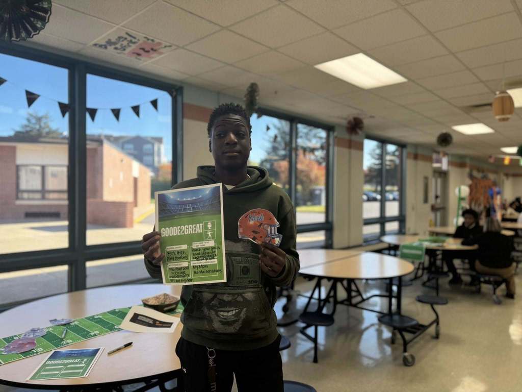 A student is standing in a school cafeteria, holding a colorful football helmet sticker that says "York" on it as well as a football-themed Goode2Great flyer. Other people can be seen in the background.