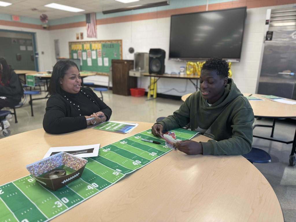 Dr. Andrea Berry-Brown, Superintendent of Schools, and a student are sitting at a round table in a school cafeteria with a green football-themed cover. The student is holding a cutout, and they appear to be engaging in a discussion. Other people can be seen in the background.