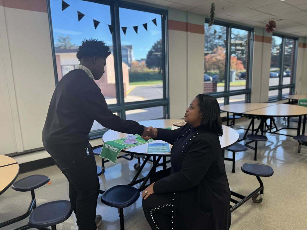 Dr. Andrea Berry-Brown, Superintendent of Schools, is sitting down shaking hands with a student who is standing up at a round table in a school cafeteria. 