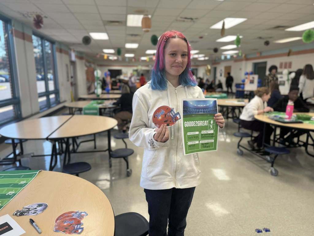 A student is standing in a school cafeteria, holding a colorful football helmet sticker that says "York" on it as well as a football-themed Goode2Great flyer. Other people can be seen in the background.
