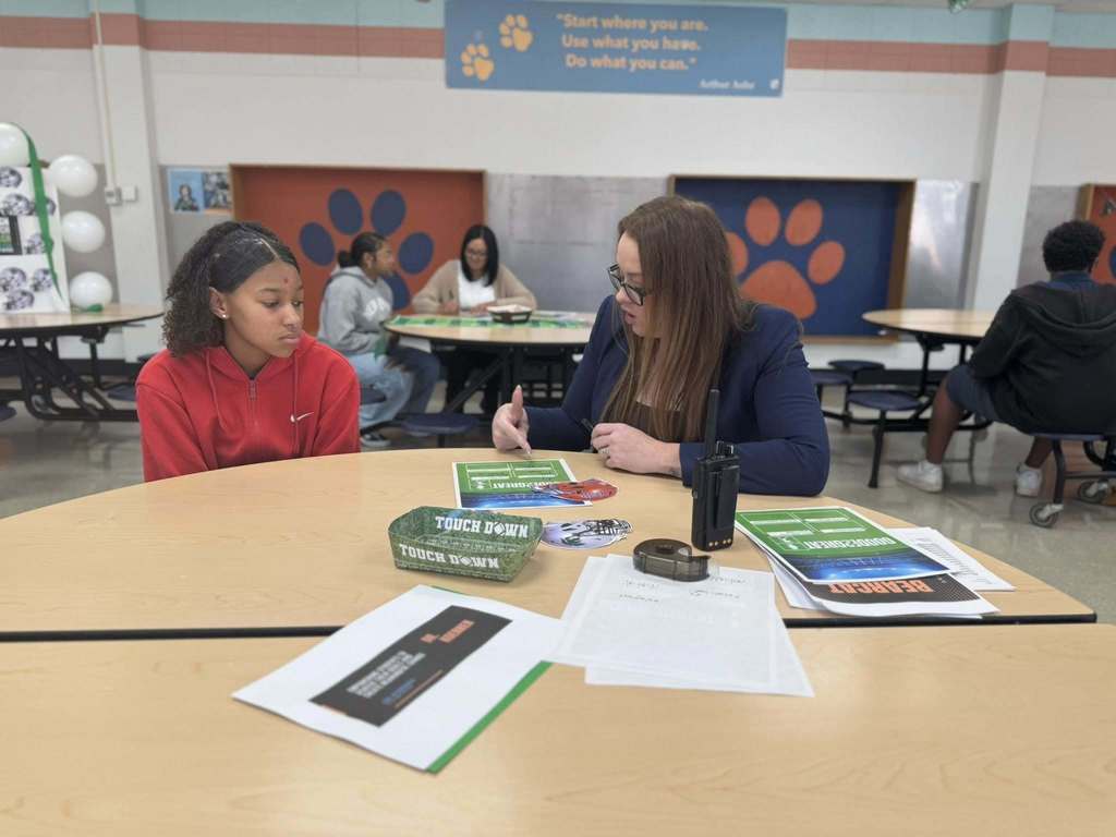 Goode PreK-8 Principal, Dr. Koerner and a student are sitting at a round table in a school cafeteria engaging in a discussion. Papers and green "Touch Down" wristbands are on the table. Other people can be seen in the background.