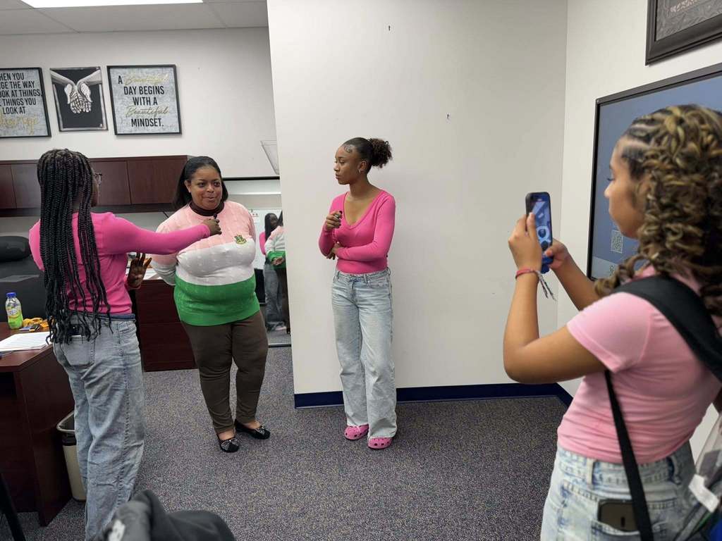 Dr. Andrea Berry-Brown, Superintendent of Schools, is engaging with three students in an office area. One of the students is holding a phone, capturing the moment, while another student is holding a microphone up to Dr. Berry-Brown and the other student is holding a microphone in her hand.