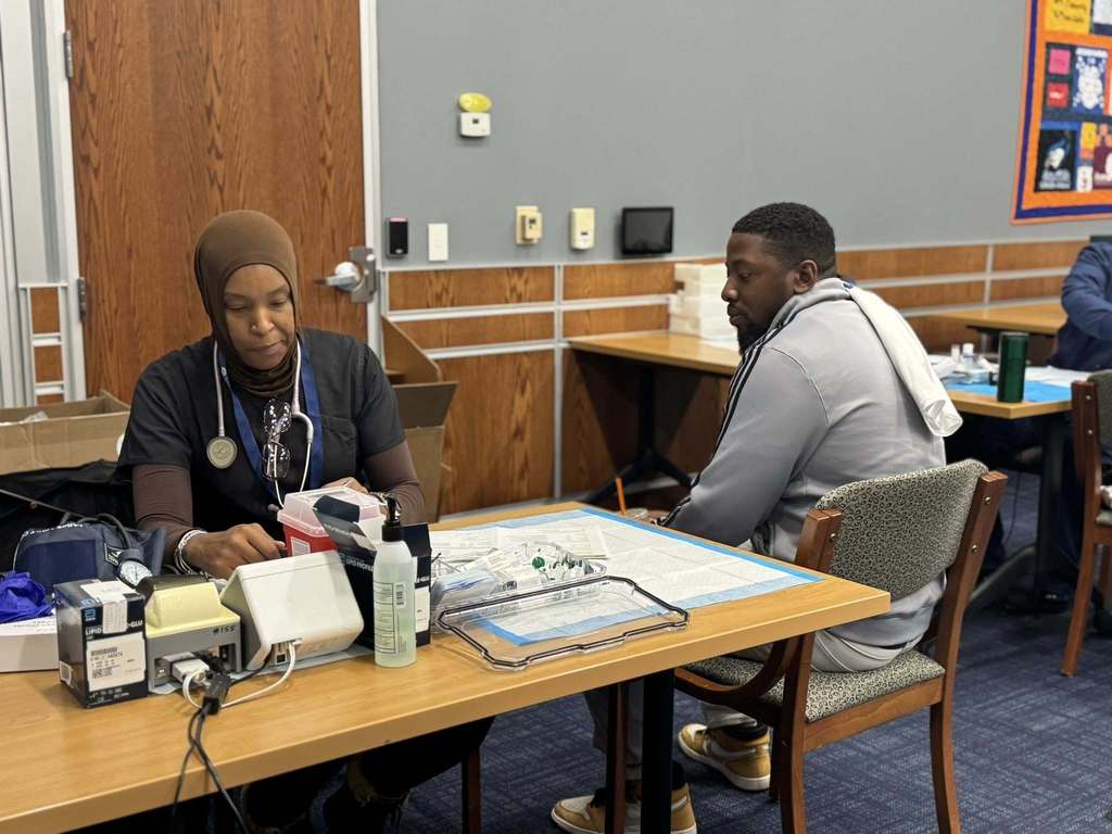 A healthcare worker wearing a stethoscope is preparing supplies at a table, while a district staff members is sitting nearby observing her.