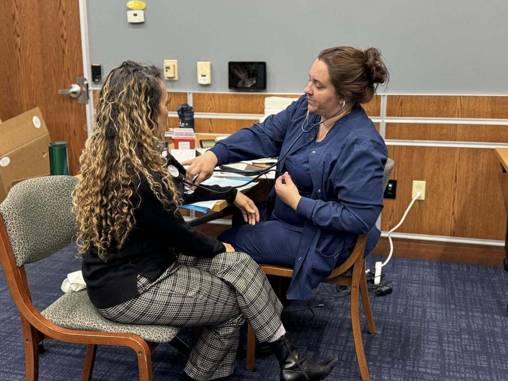 A healthcare professional in blue scrubs is checking a district staff member's blood pressure in a conference room setting.