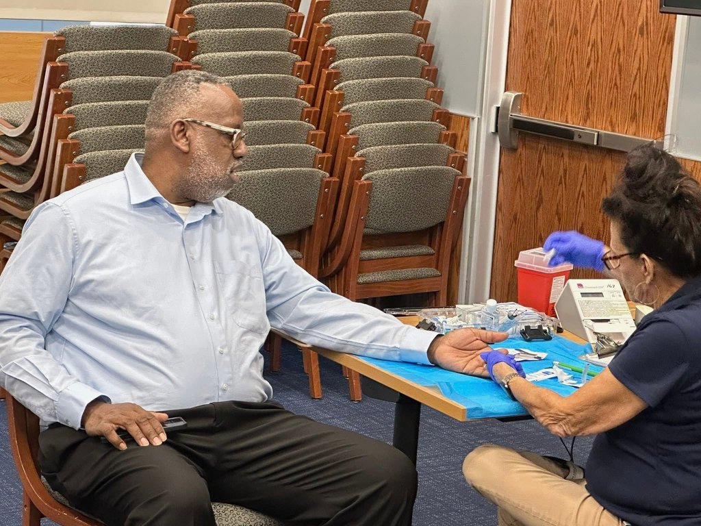 A district staff member is sitting in a conference room setting receiving a medical test. A healthcare worker in gloves is preparing his arm. Stacked chairs are in the background.