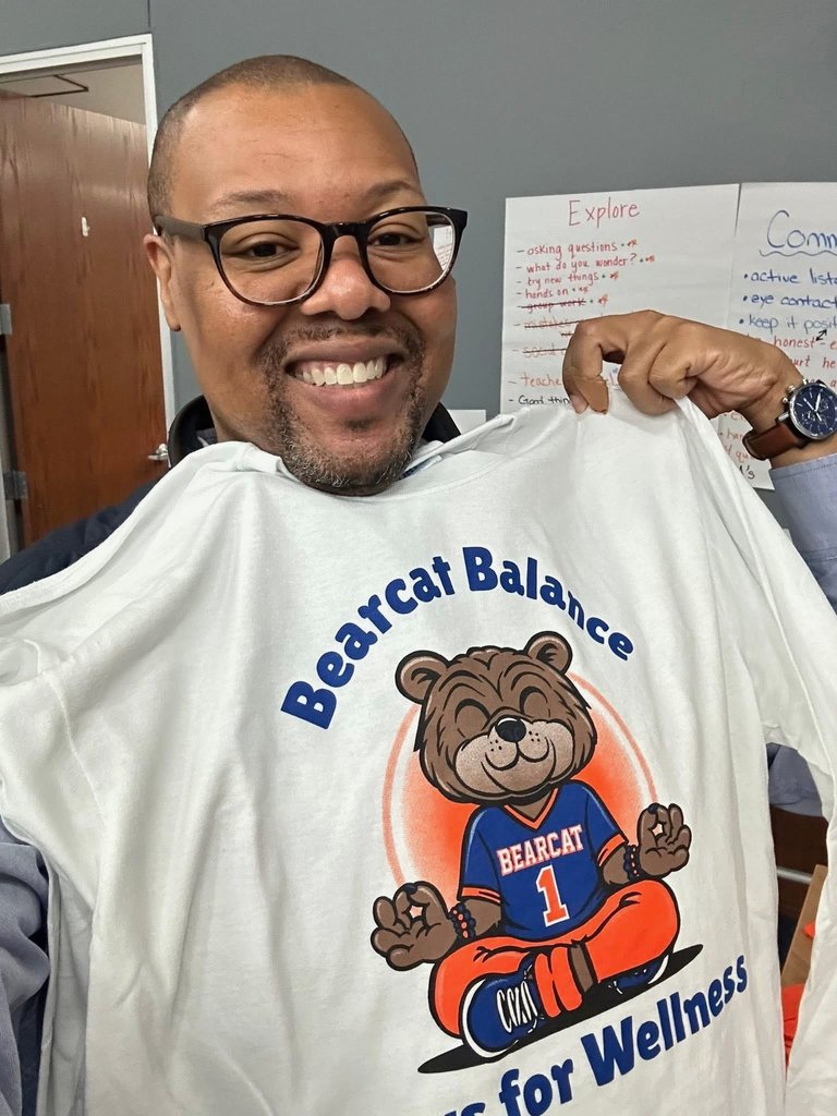 A district staff member is holding up a white shirt with a cheerful cartoon bear that is meditating. The shirt reads "Bearcat Balance - Paws for Wellness". There are two charts on the wall in the background.