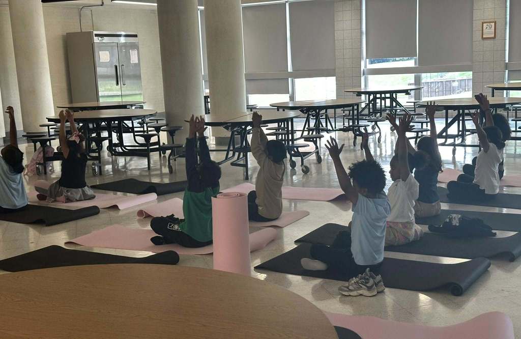 A group of students are sitting on black and pink mats practicing yoga poses in a school cafeteria. Their arms are stretched in the air.