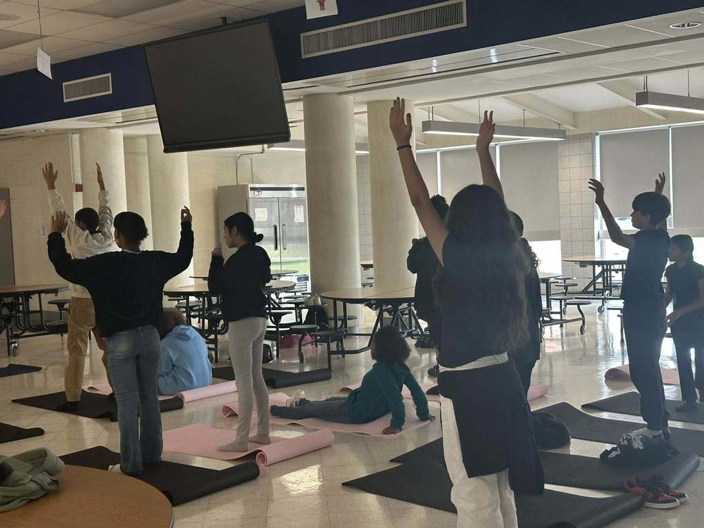 A group of students are practicing yoga poses on black and pink mats in a school cafeteria. Their arms are stretched in the air.
