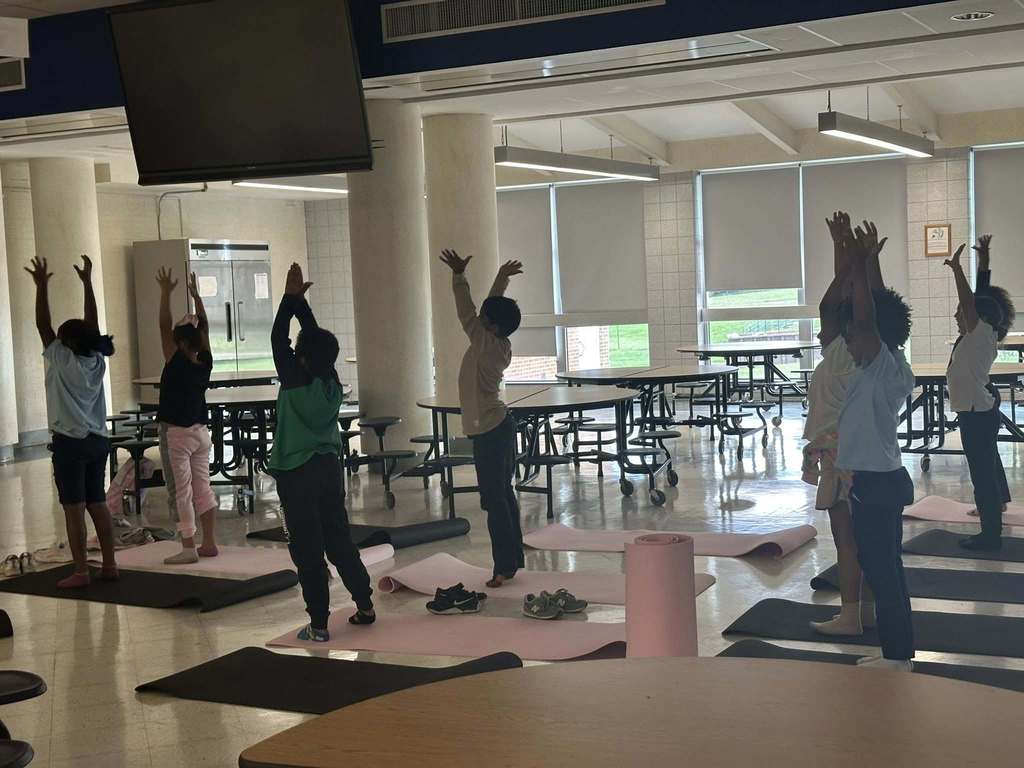 A group of students are practicing yoga poses on black and pink mats in a school cafeteria. Their arms are stretched in the air.