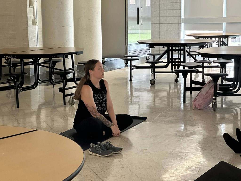 A yoga instructor is sitting cross-legged on a black mat in a school cafeteria.