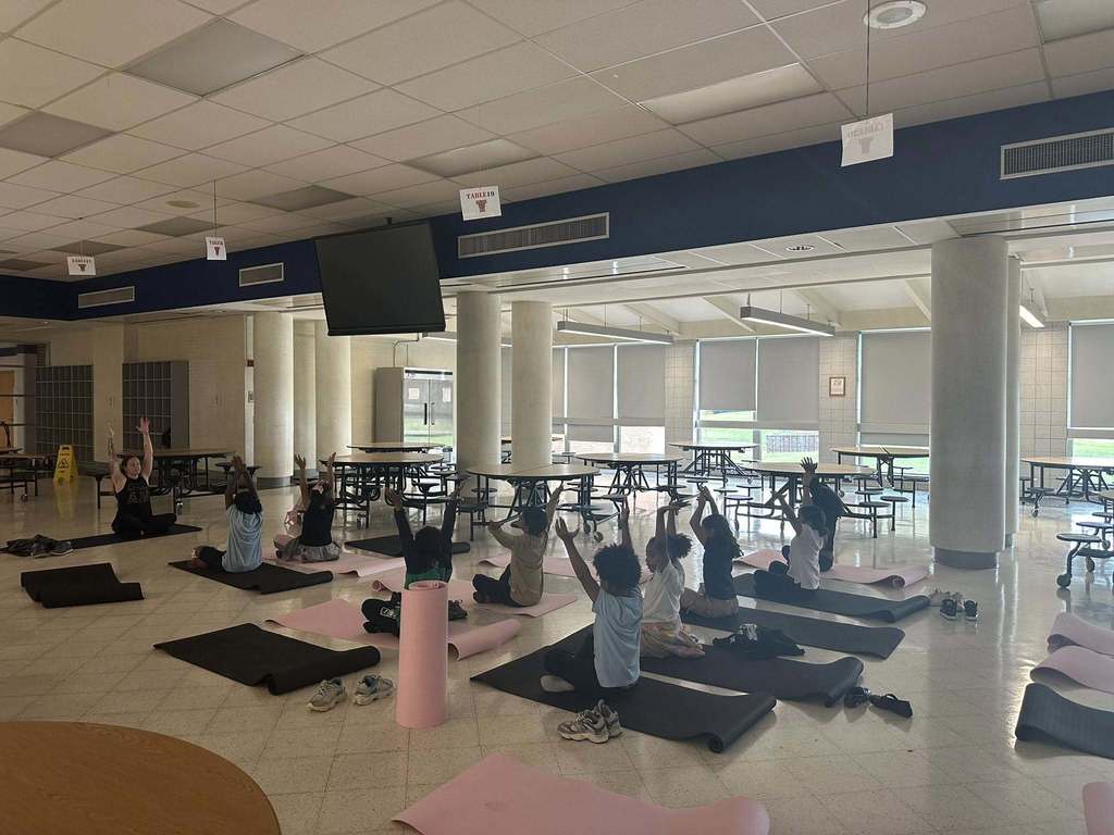 A group of students, following a yoga instructor, are practicing yoga poses on black and pink mats in a school cafeteria. Their arms are stretched in the air.