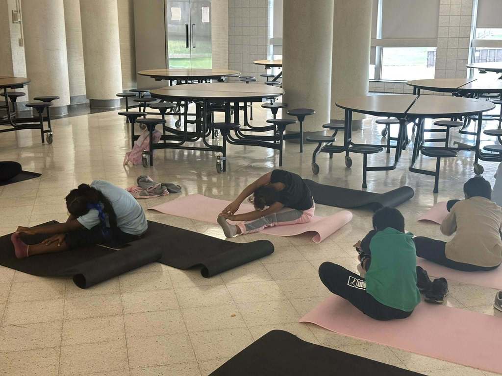 A group of students are practicing yoga poses on black and pink mats in a school cafeteria.