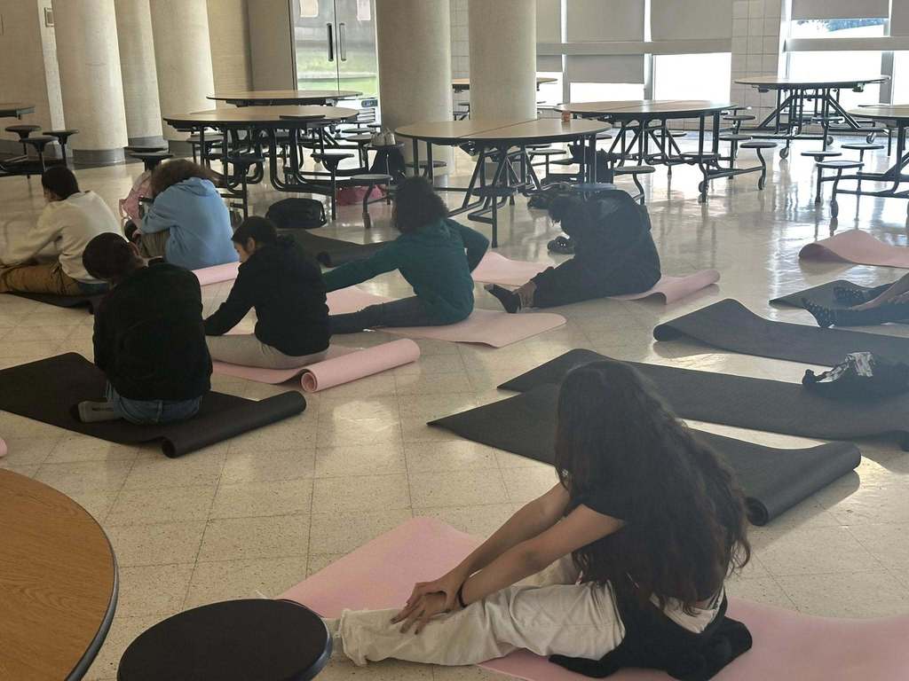 A group of students are practicing yoga poses on black and pink mats in a school cafeteria.