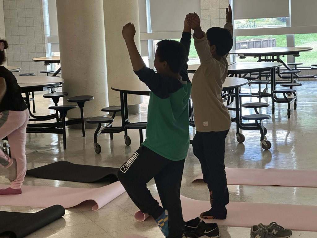 Two students are practicing yoga poses on mats inside of a school cafeteria. Their arms are stretched in the air.