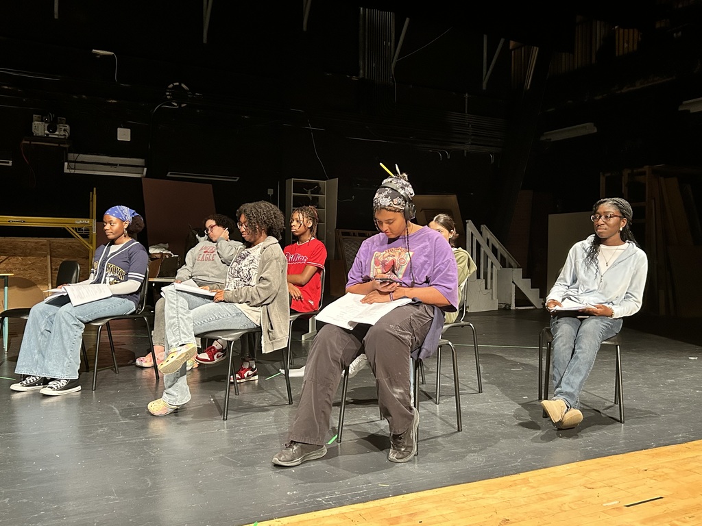 A group of seven students are sitting on chairs in a school auditorium rehearsing on a dimly lit stage.