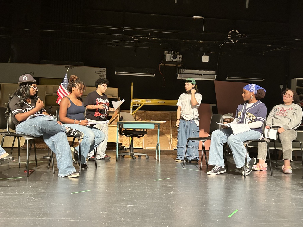 A group of six students are sitting at chairs rehearsing on a dimly lit stage in a school auditorium. A desk is nearby and an American flag is also in the background. 