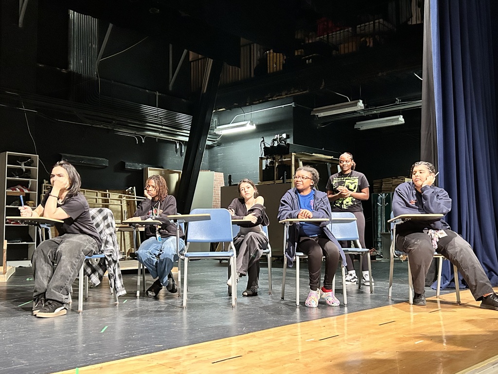 A group of six students are rehearsing on a dimly lit stage in a school auditorium. Five of the students are sitting down at desks, while the other student is standing up near them.
