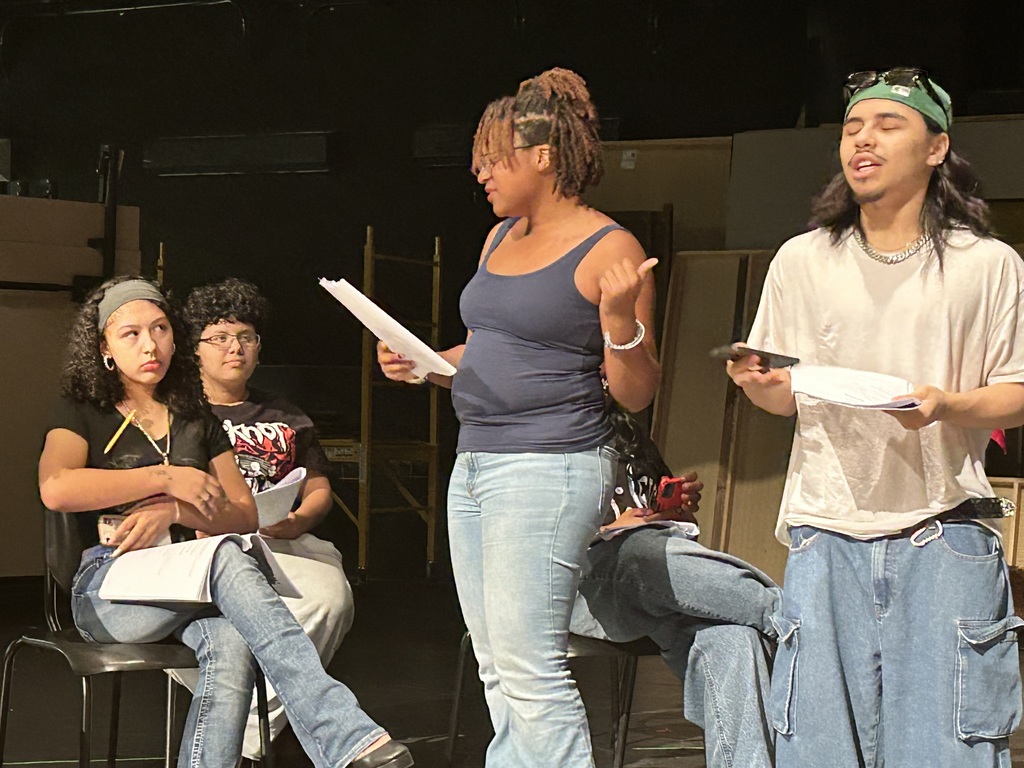 A group of four students are rehearsing in a school auditorium. Each of the students are holding scripts. One student is speaking passionately while other student are listening attentively on a dimly lit stage, suggesting a theatrical setting. Another student can be seen in the background.