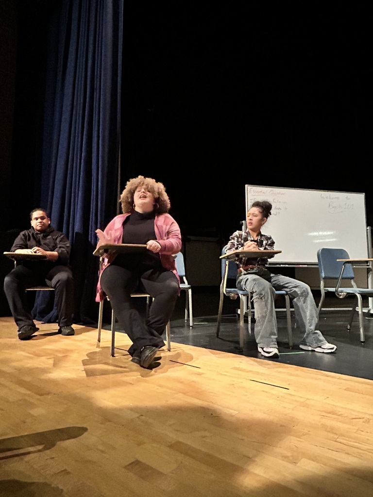 A group of three students are sitting at desks rehearsing in a school auditorium. One of the students appears to be singing, while the other two students are observing. A whiteboard can be seen in the background.