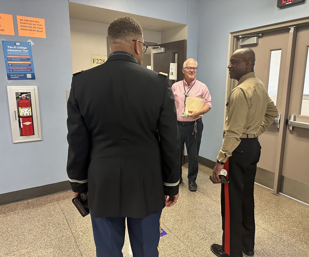 Three men are conversing in a school hallway near fire safety signs and closed doors. One of the men is wearing a black uniform, another man is wearing a tan shirt, and the third man is holding documents in his hand.