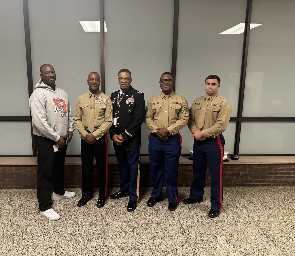 A group of five men, including two district staff members and military personnel in uniform, are standing together against a glass wall backdrop.