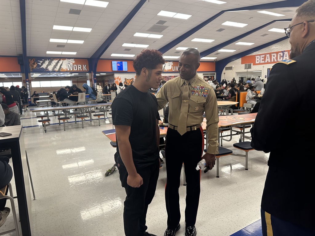 A young student and a uniformed officer are conversing in a school cafeteria filled with students sitting at tables, promoting military engagement.