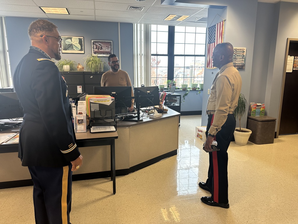 Two men dressed in military uniforms are conversing with a district staff member at a desk in an office setting with large windows and American flag.