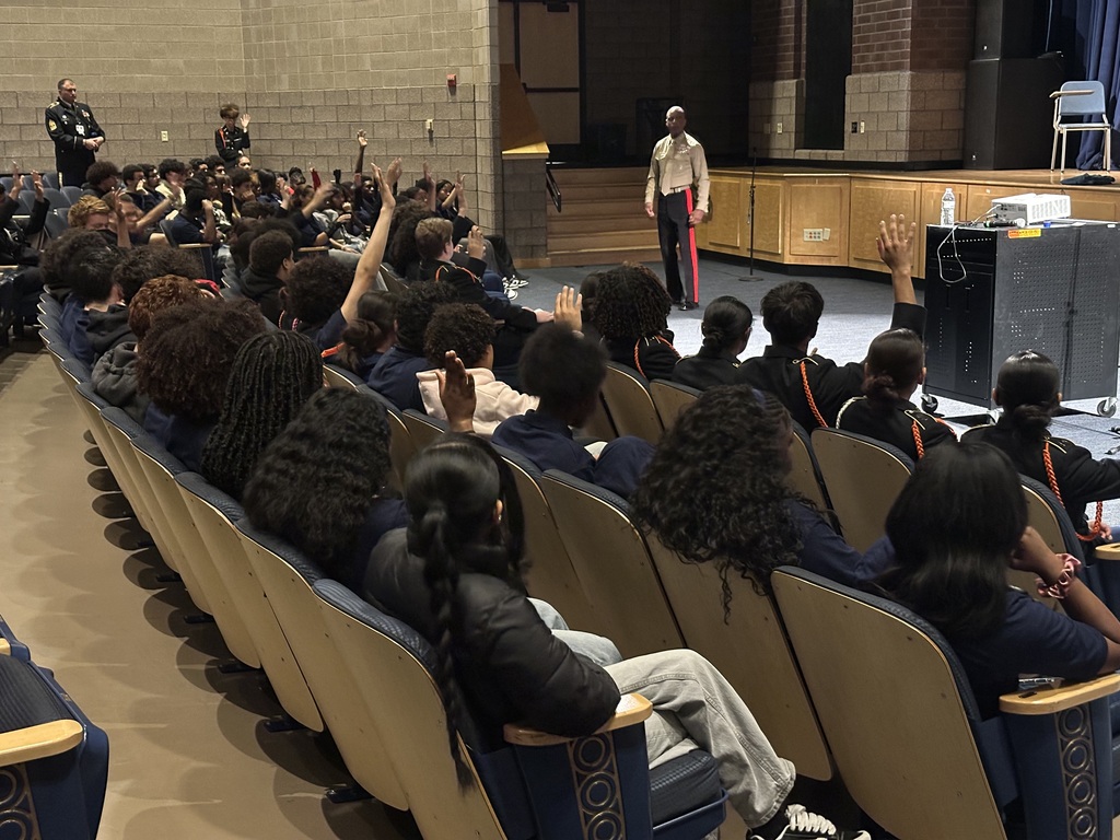 A uniformed officer is standing in front of an attentive audience, addressing a large group of students who are seated in a school auditorium.