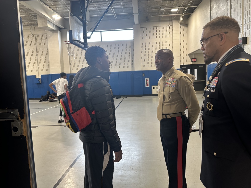Two military officials are conversing with a young student in a school gym, while other students are playing basketball in the background.