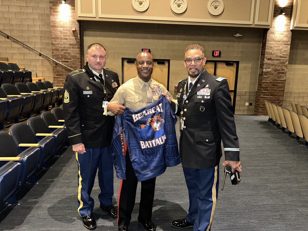 Three uniformed military officers are standing together in a school auditorium, smiling for a photo. The uniformed officer in the center is holding a blue jacket that reads “BEARCAT BATTALION U.S. ARMY” with a growling bearcat mascot printed on it. 