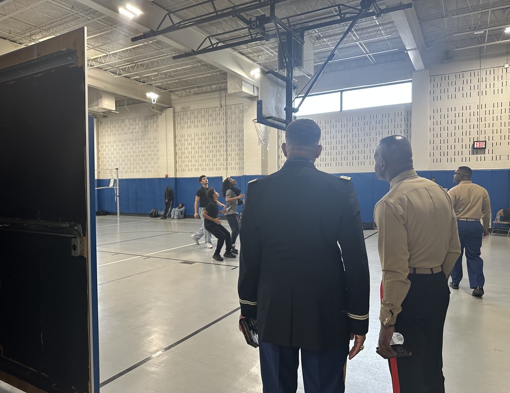 Two military personnel are watching a group of students play a basketball game in a school gym, with other students sitting down nearby in the background.