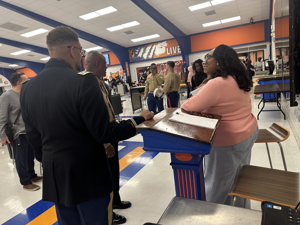 A group of adults, including district staff members and military personnel, are engaging in conversation in a school cafeteria.