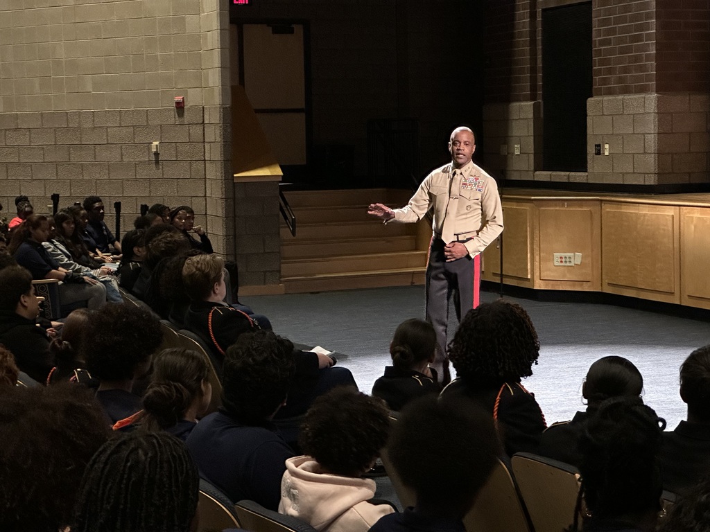 A speaker in military uniform is addressing an attentive audience in a school auditorium, engaging with students during a presentation.