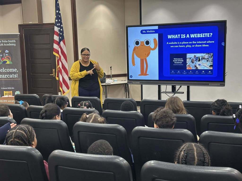 Supervisor of Communications and Marketing, ShaiQuana Mitchell is presenting to a group of seated students, explaining a digital slide titled "What is a Website?" featuring an orange cartoon character in a Large Group Instruction (LGI) room.