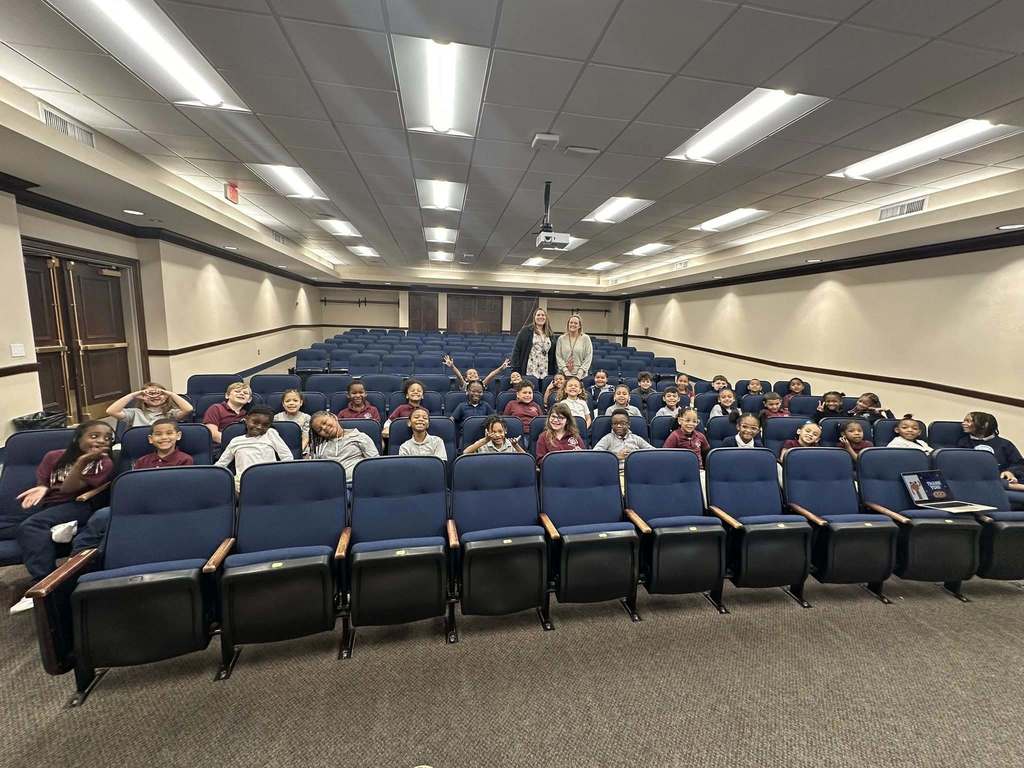 A diverse group of students are sitting in a Large Group Instruction (LGI) room with blue seats, smiling and waving at the camera, creating a lively atmosphere. Two teachers are standing behind the students also smiling at the camera. 
