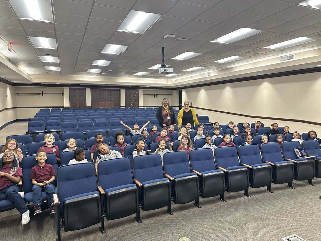 A group of smiling students and two district staff members are posing in a Large Group Instruction (LGI) room. The students are sitting in blue seats, displaying excitement and joy.