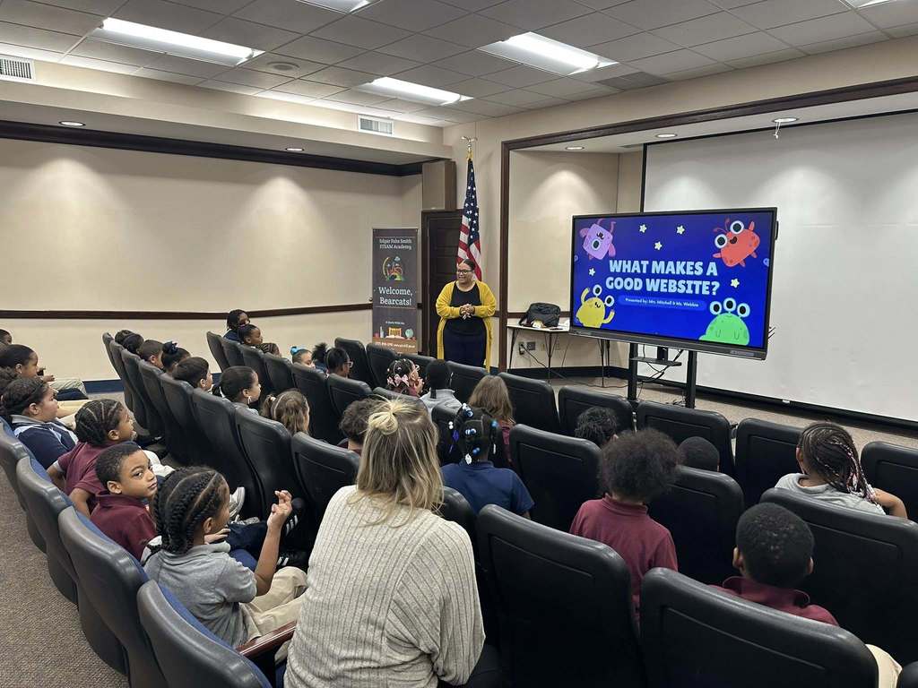 Supervisor of Communications and Marketing, ShaiQuana Mitchell is presenting to a group of attentive students in a Large Group Instruction (LGI) room, discussing "What Makes a Good Website" on a screen with colorful monster graphics. 