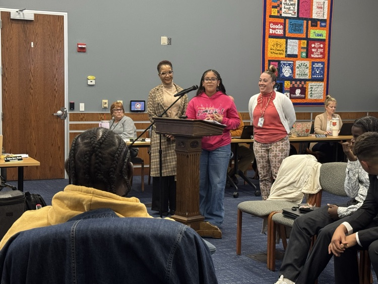 A student speaking at a podium in a conference room surrounded by district Administration, School Board members and the community. 