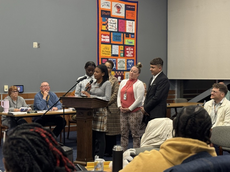A student speaking at a podium in a conference room surrounded by district Administration, School Board members and the community. 