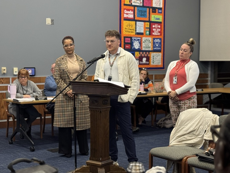 A district staff member speaking at a podium in a conference room surrounded by district Administration, School Board members and the community. 