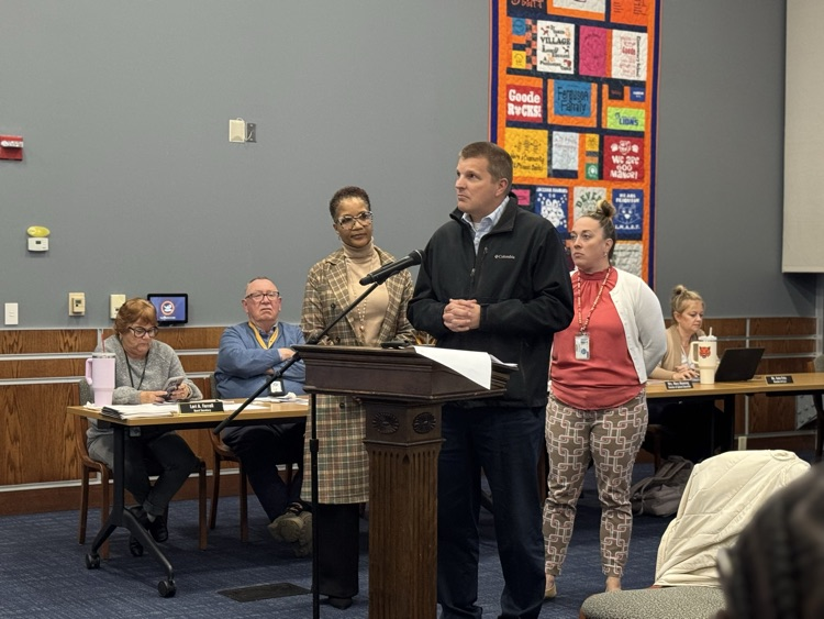 A district staff member speaking at a podium in a conference room surrounded by district Administration, School Board members and the community. 