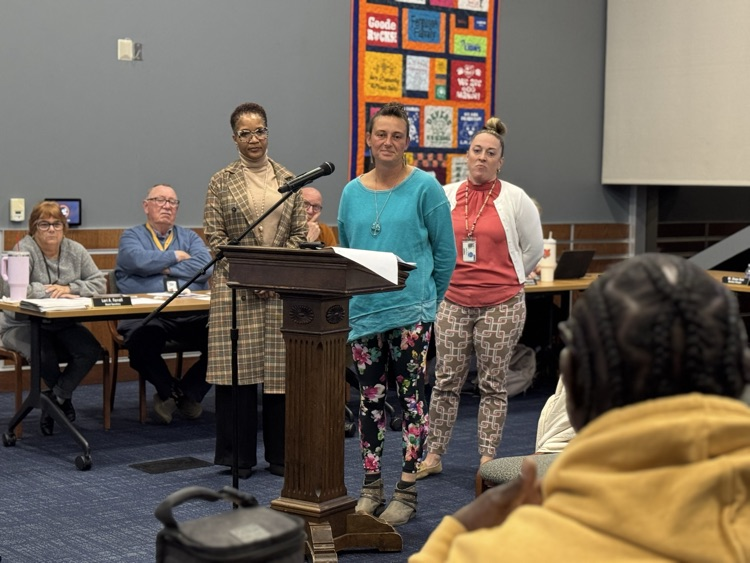 A district staff member speaking at a podium in a conference room surrounded by district Administration, School Board members and the community. 