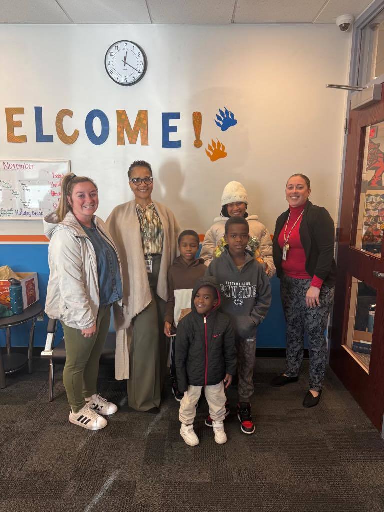 A group of four adults, including district staff members and four children are standing in front of a "Welcome!" sign on a wall. One of the adults is holding a fruit basket in her hand. 