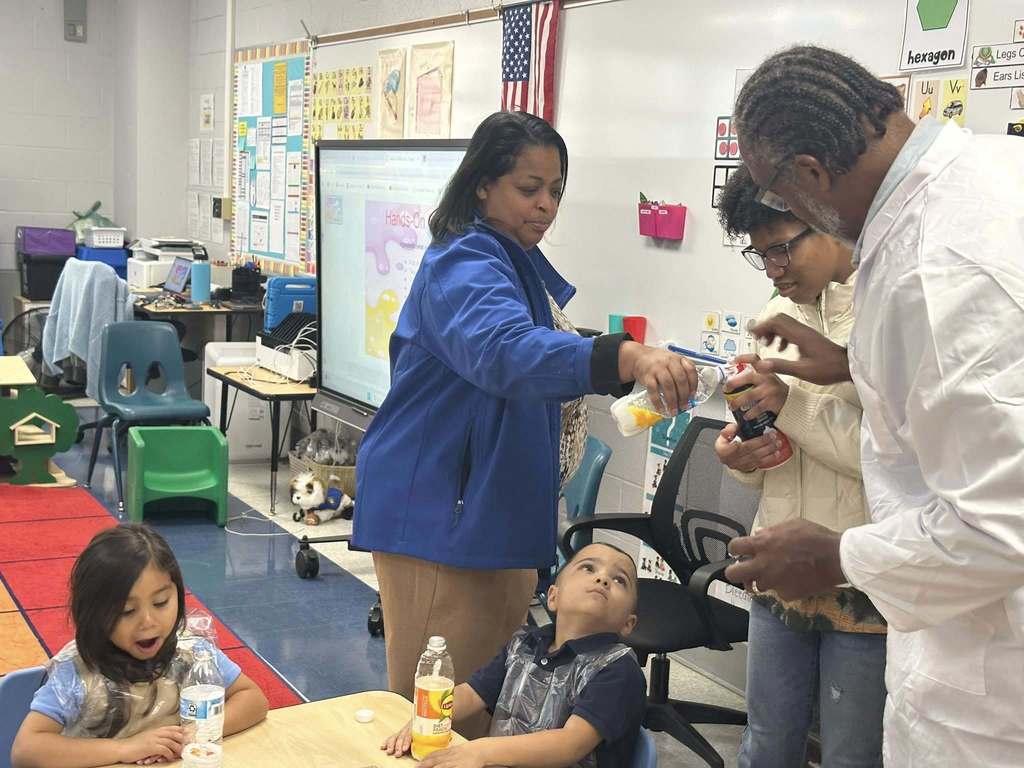 Dr. Andrea Berry-Brown, Superintendent of Schools, a district staff member and a student are assisting two young students with a science experiment using bottles in a classroom. A screen is displaying colorful graphics. Posters can also be seen hanging on the walls.