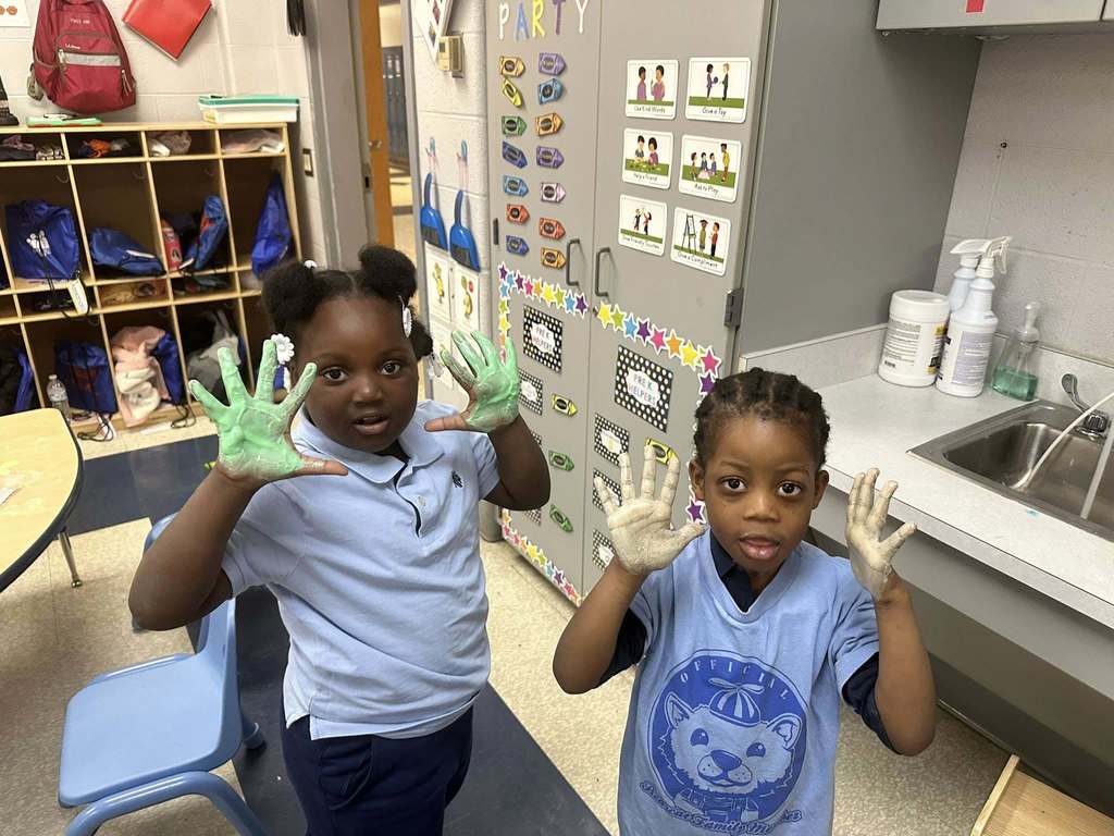 Two young students are holding up both of their hands that are covered in slime in a classroom. 