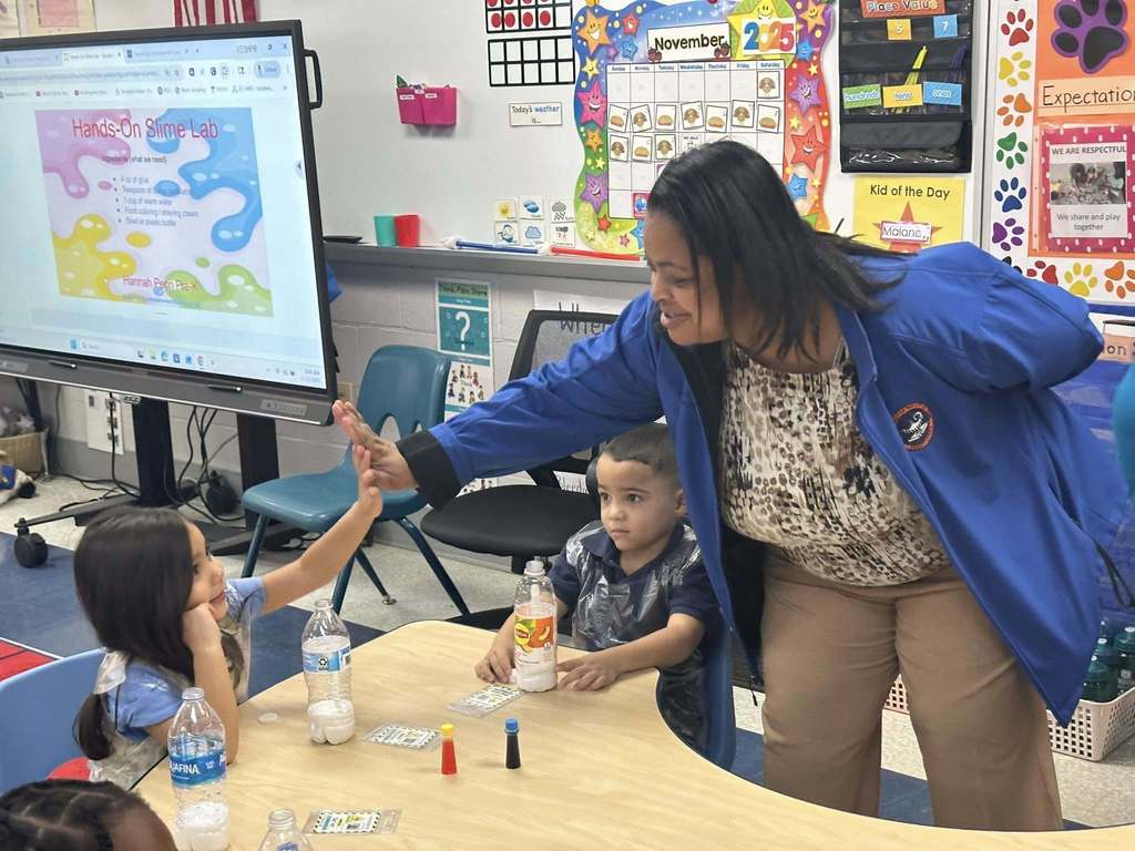 Dr. Andrea Berry-Brown, Superintendent of Schools, is giving a high-five to a young student who is sitting at a table in a classroom. Another student is sitting nearby observing. Posters can be seen hanging on the walls as well as a projector screen.