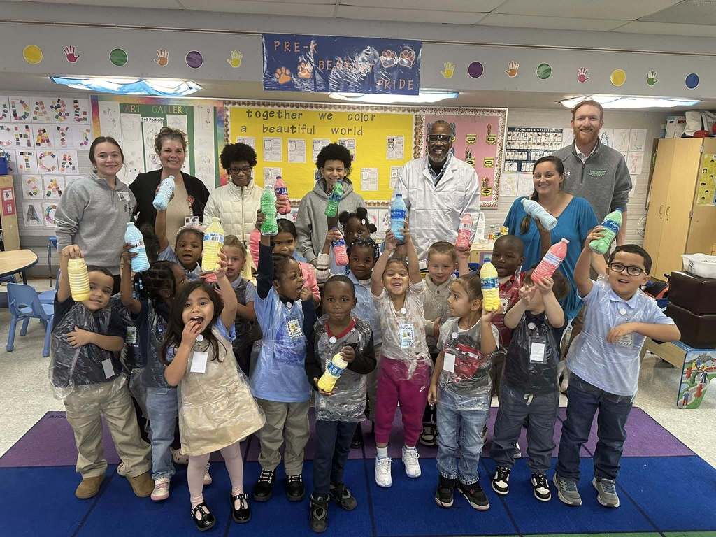 A diverse group of young students and district staff members are standing on a colorful carpet in a classroom. The students are holding up vibrant, painted bottles.
