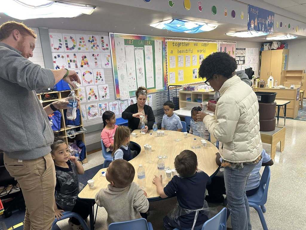 A group of young students are sitting around a table engaging in a classroom experiment. A district staff member and an older student are assisting the younger students. Posters can be seen hanging on the walls.