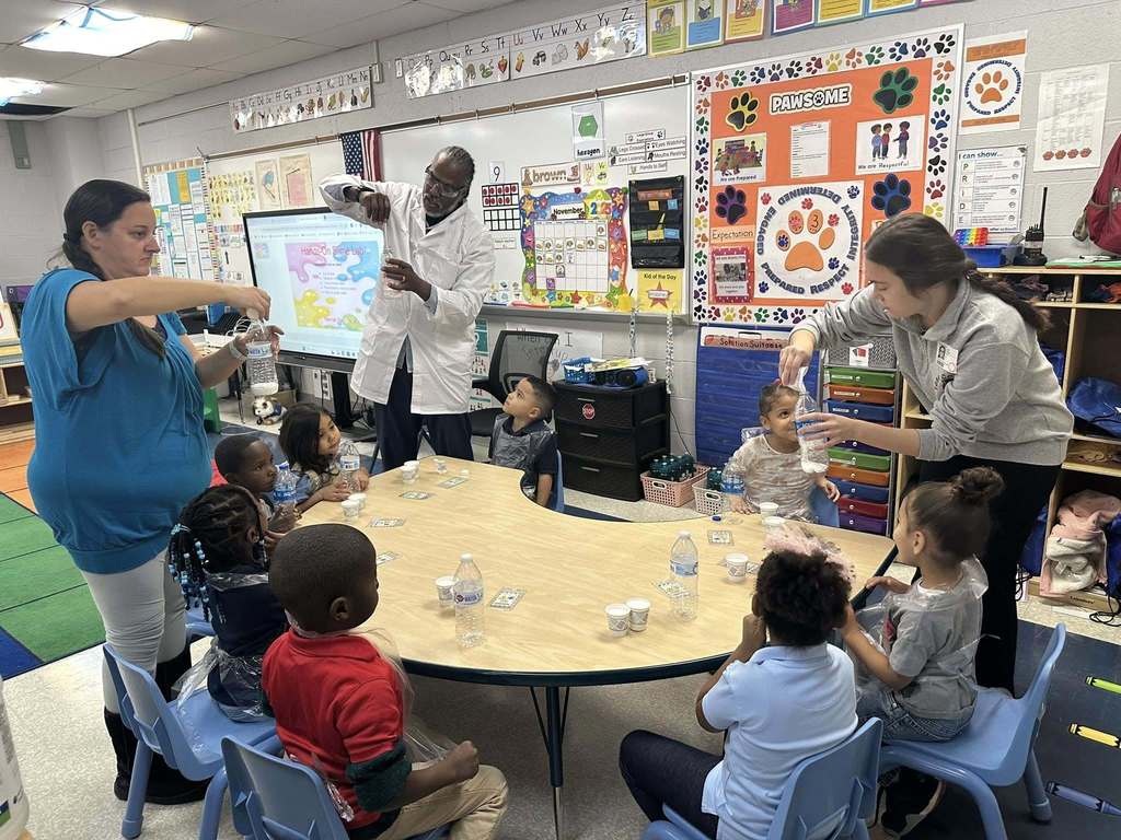 A group of young students are sitting around a table engaging in a classroom experiment. Three district staff members are assisting the younger students. Posters can be seen hanging on the walls as well as a projector screen.
